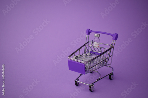 Toy shopping basket with glass bubbles. In basket a vial with a sticker of a vaccine against covid. On a purple background.