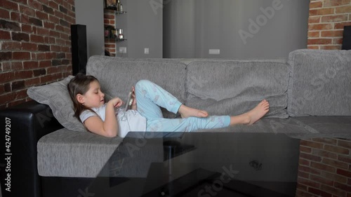 A little girl lies at home on the couch and looks into a smartphone. The child plays and watches the video. The girl has bare feet. The child is wearing pajamas. 4K