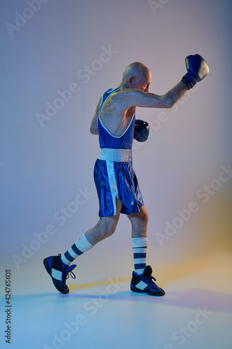 Senior man wearing sportwear boxing isolated on gradient studio background in neon light. Concept of sport, activity, movement, wellbeing. Copyspace, ad.