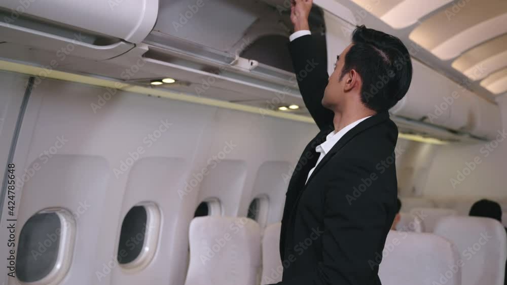 Businessman puts a suitcase in a baggage compartment Flight attendant welcome passenger on board, advice seat and bring luggage to store in overhead bin compartment in business class in airplane cabin Businessman puts a suitcase in a baggage compartment Flight attendant welcome passenger on board, advice seat and bring luggage to store in overhead bin compartment in business class in airplane cabin