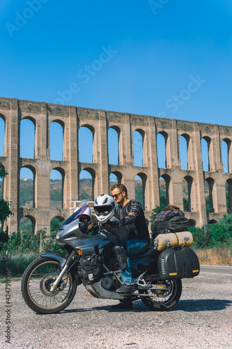 A man in a protective motorcycle outfit poses against the backgrond of an old Aqueduct of Vanvitelli, Caroline. Sunny day. Glasses and turtle jacket. Motorbikers and travel. Vertical photo