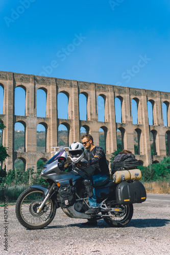 A man in a protective motorcycle outfit poses against the backgrond of an old Aqueduct of Vanvitelli, Caroline. Sunny day. Glasses and turtle jacket. Motorbikers and travel. Vertical photo photo