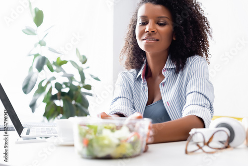 Smiling african american woman taking takeaway salad near laptop on blurred background