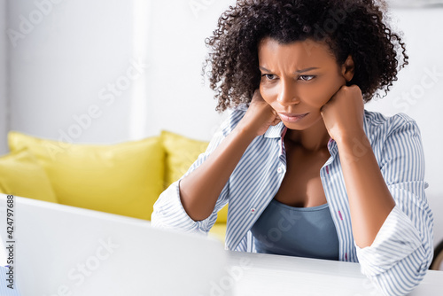 Concentrated african american freelancer looking at laptop on blurred foreground at home