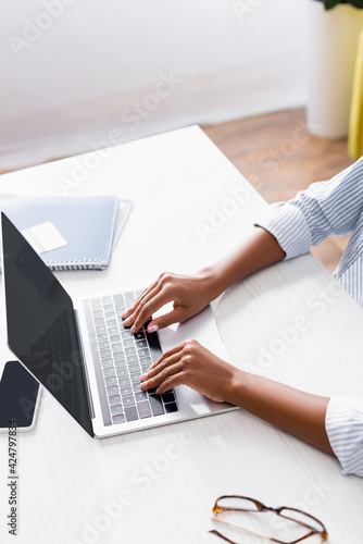 Cropped view of african american teleworker using laptop near eyeglasses and notebook on blurred background