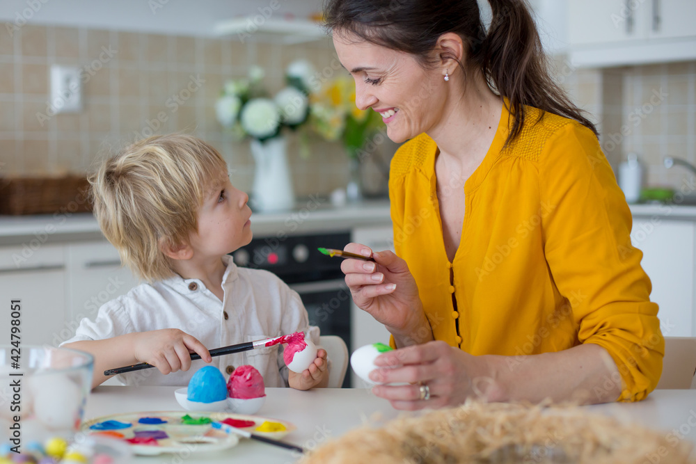 Obraz premium Beautiful blond child, toddler boy, painting easter eggs with mother at home, making easter wreath