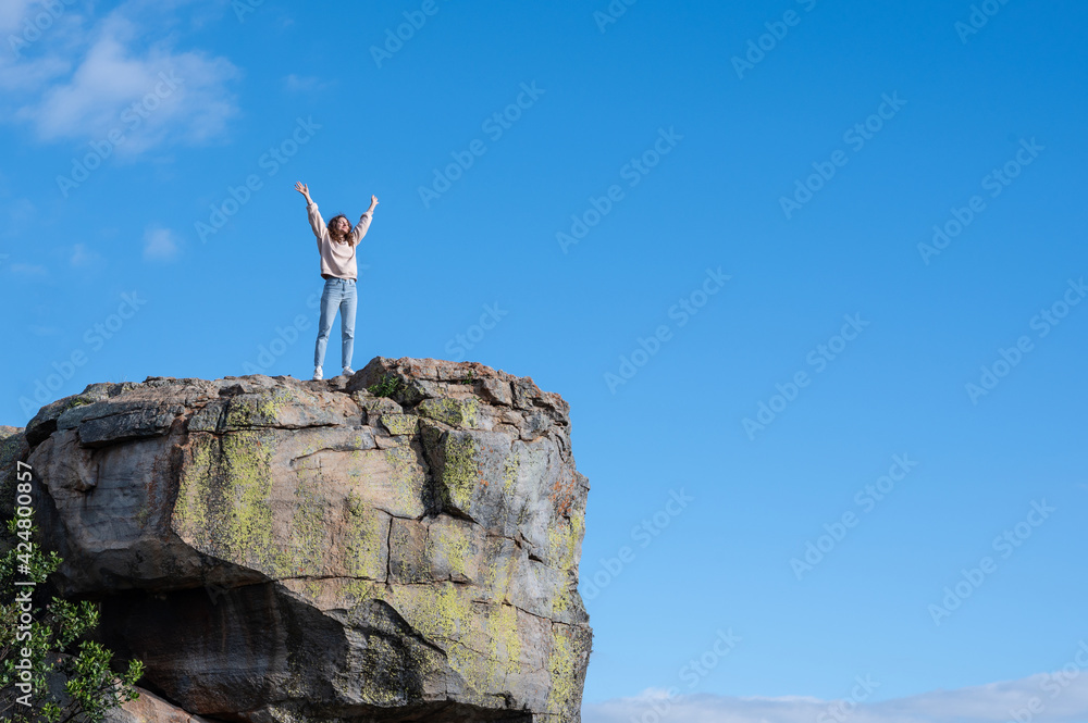 Woman standing on the edge of the rock and raising hands against blue sky Woman standing on the edge of the rock and raising hands against blue sky