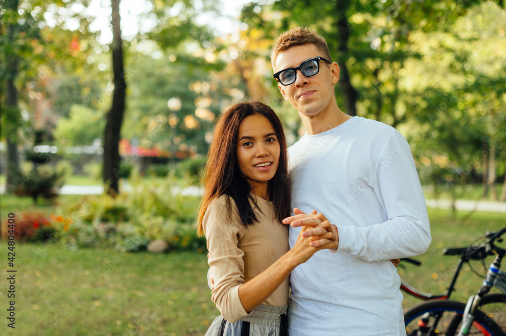 Fototapeta premium Attractive cute couple is standing in the park, posing for the camera, holding their hands and smiling. A caucasian man and hispanic woman are dating and feeling happy.
