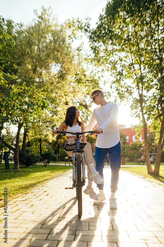 Obraz premium Vertical photo of two people, a couple, in the city park on a sunny day, a woman is sitting on a bicycle, a man is holding her and talking to her. Lifestyle concept.