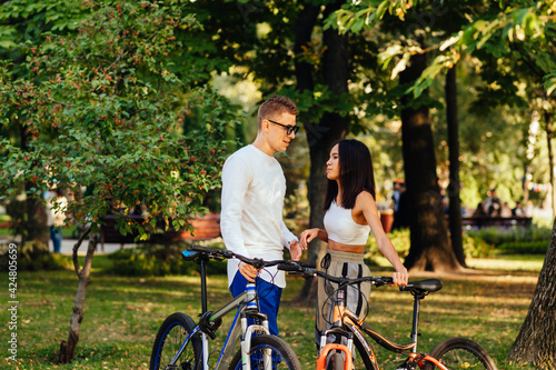 Youthful couple of male and female bicyclists is having a thoughtful conversation in the summer park. Boyfriend and girlfriend are having a perfect date riding their bikes.