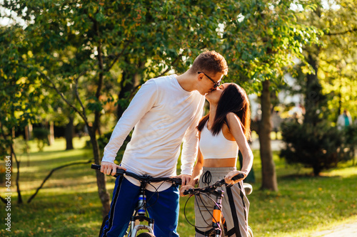 Photo of a lovely kissing couple standing outside with their bicycles, feeling romantic in the city park on a sunny summer day. Relationship concept.
