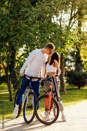 Vertical photo of a lovely kissing couple standing outside with their bicycles, feeling romantic in the city park on a sunny summer day. Relationship concept.