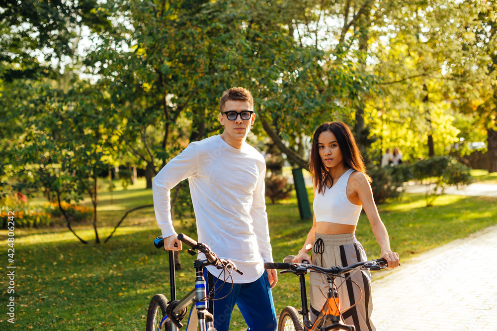 Obraz premium Handsome caucasian man and pretty hispanic woman are standing in the park, holding their bikes, posing for the photo and looking at the camera. Lifestyle, cycling concept.