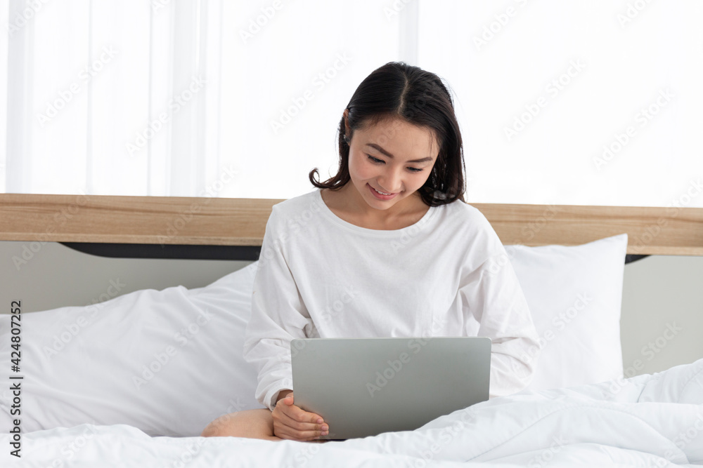 Young female sitting on the bed and working on a laptop. Young woman working online from home with laptop on her bed. Asian woman using notebook computer at home and sitting on her bed. Young female sitting on the bed and working on a laptop. Young woman working online from home with laptop on her bed. Asian woman using notebook computer at home and sitting on her bed.