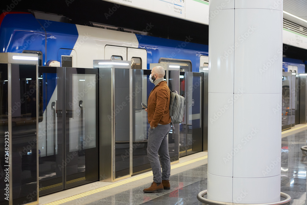 A man in a medical face mask to avoid the spread of coronavirus is holding a cellphone while waiting for a modern train at the subway platform. A bald guy in a surgical mask is keeping social distance A man in a medical face mask to avoid the spread of coronavirus is holding a cellphone while waiting for a modern train at the subway platform. A bald guy in a surgical mask is keeping social distance
