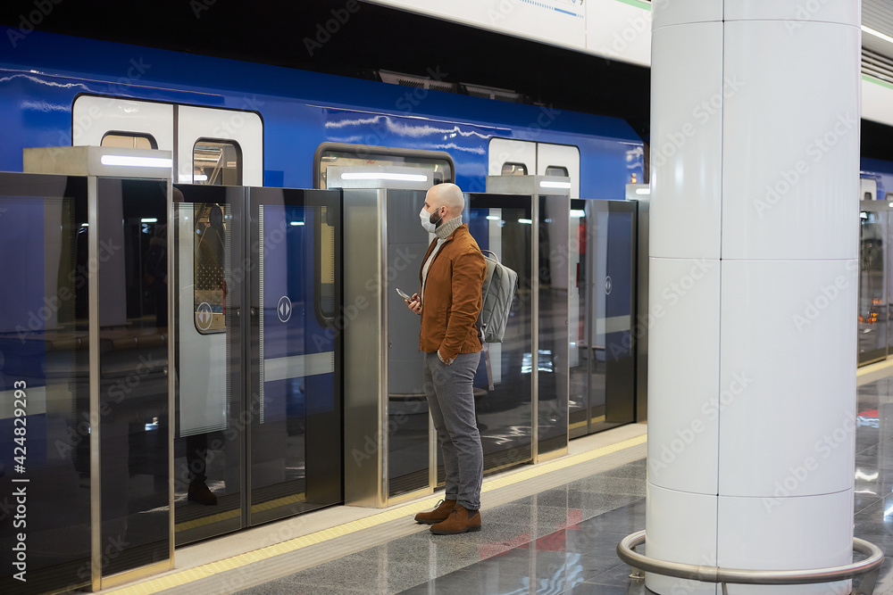 A man in a face mask to avoid the spread of coronavirus is holding a smartphone while waiting for a modern train at the subway platform. A bald guy in a surgical mask is keeping social distance. A man in a face mask to avoid the spread of coronavirus is holding a smartphone while waiting for a modern train at the subway platform. A bald guy in a surgical mask is keeping social distance.
