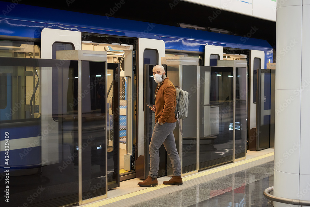 A man in a medical face mask to avoid the spread of coronavirus is holding a smartphone while entering the modern subway car. A bald guy in a surgical mask is keeping social distance. A man in a medical face mask to avoid the spread of coronavirus is holding a smartphone while entering the modern subway car. A bald guy in a surgical mask is keeping social distance.