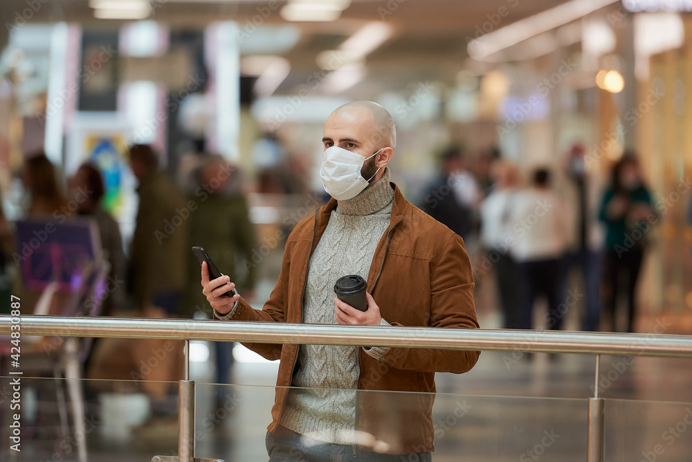 A man with a beard in a face mask is looking to the left and holding a smartphone and a cup of coffee in the shopping center. A bald guy in a surgical mask is keeping social distance.