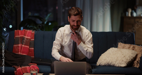 Young caucasian smiling man returning home opening laptop to play favorite video game loosening necktie enjoying leisure evening alone.