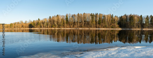 Panoramic view of the blue river. The near shore is covered with drifts of ice, the far shore is with trees and bushes without foliage. Spring evening with clear skies and sunshine.