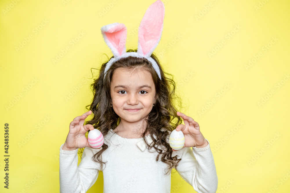 Happy curly little girl wears bunny ears, picks up colored Easter eggs, isolated on yellow background. Easter concept