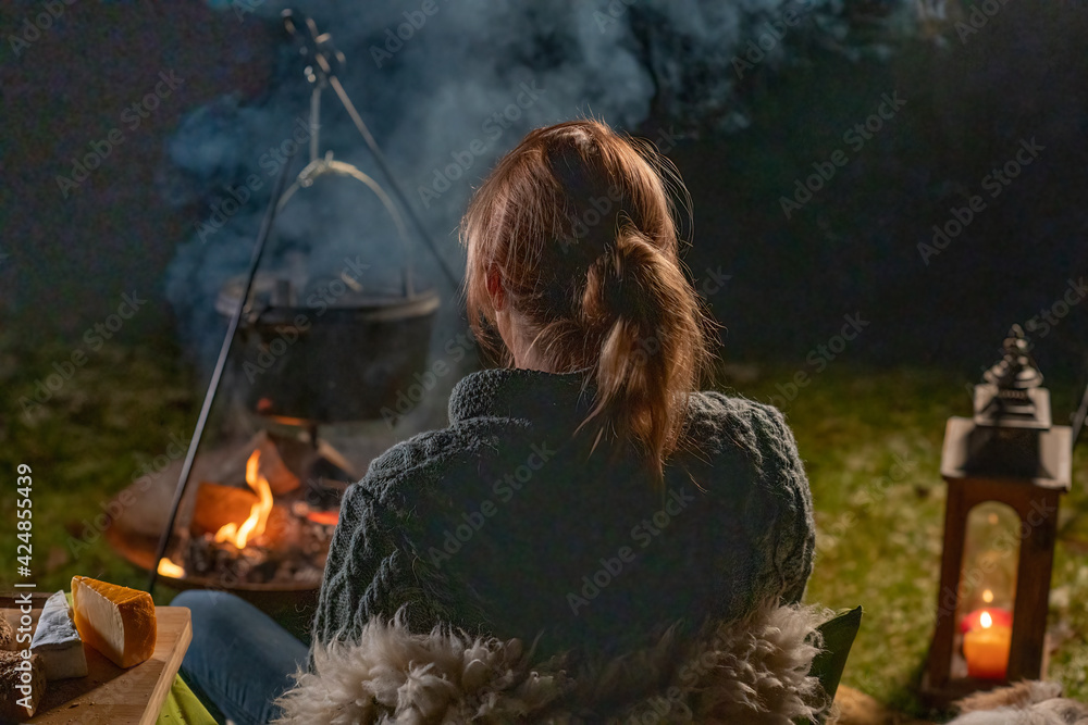 Fototapeta premium Young blond woman sits outside by the campfire and latern in the woods. It is dark in winter and she is warm by the fire in the forest. In half body, seen from behind