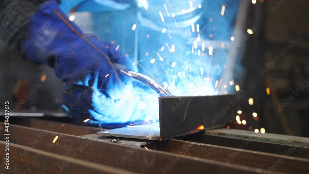 Welder in a protective mask and gloves weld metal parts on a special table using a welding machine. Close-up of the welding process of metal parts in the workshop. Welder in a protective mask and gloves weld metal parts on a special table using a welding machine. Close-up of the welding process of metal parts in the workshop.
