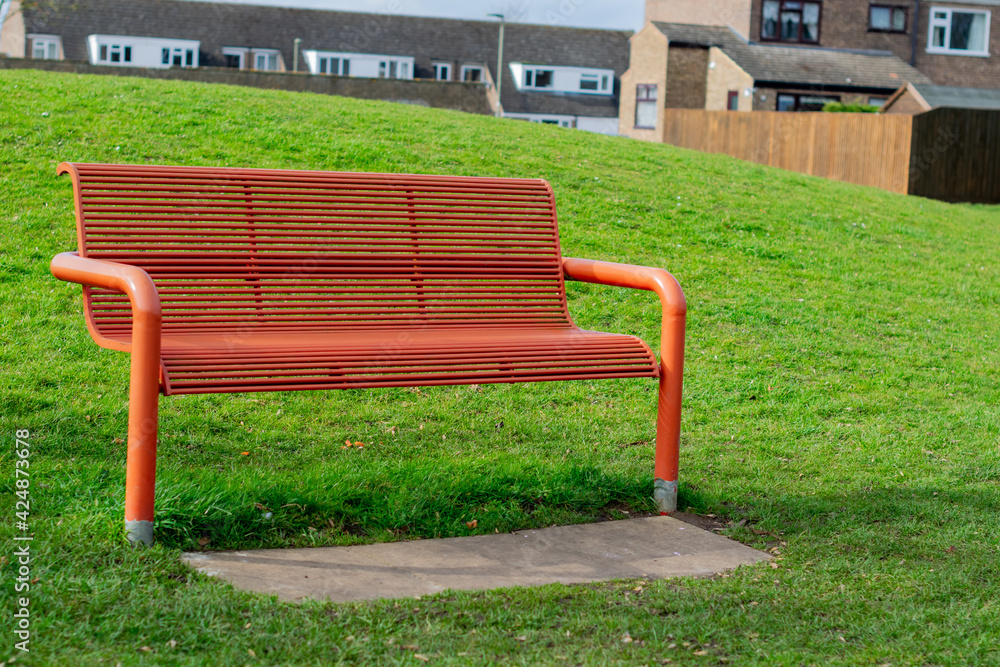 Metal orange bench in the park surrounded with bright green grass with british houses in the background, beautiful day in the community park, single empty metal bench