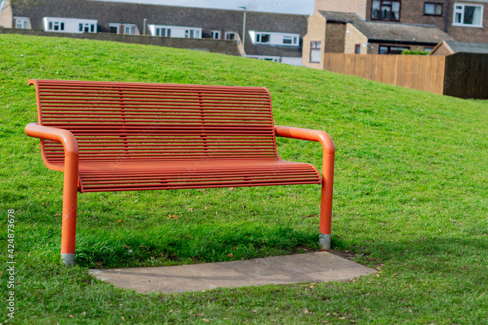 Metal orange bench in the park surrounded with bright green grass with british houses in the background, beautiful day in the community park, single empty metal bench Metal orange bench in the park surrounded with bright green grass with british houses in the background, beautiful day in the community park, single empty metal bench