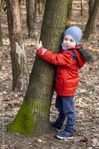 A cute little boy wearing an orange jacket  blue pants and a cap  cuddling up to a tree covered with moss. In the background  other trees and dry leaves lying on the ground.