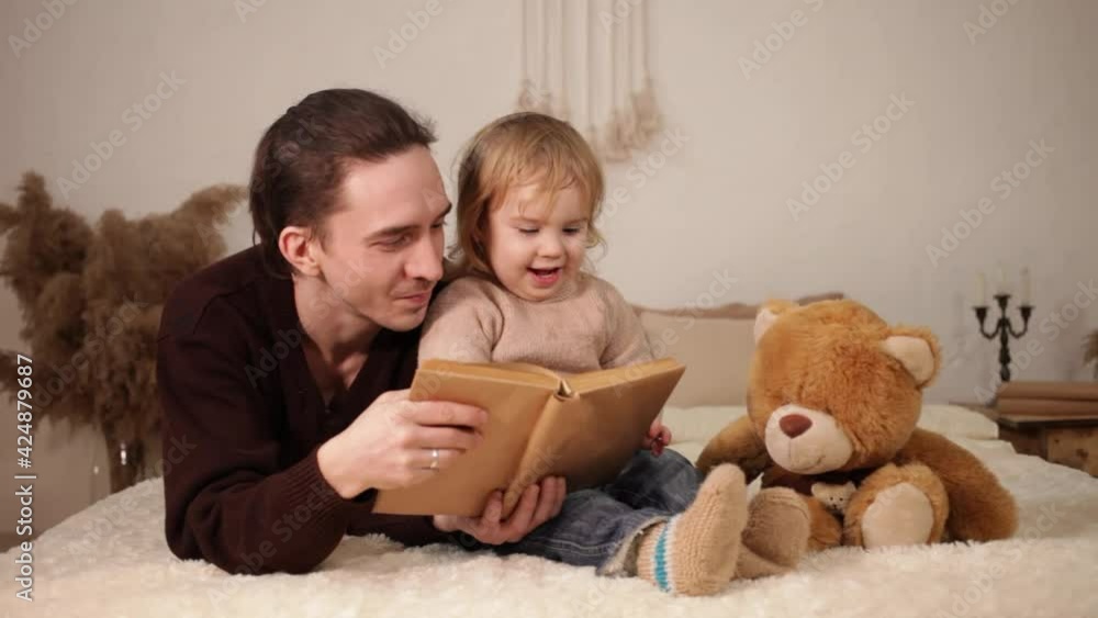 A young dad reads his daughter a book on a bed in the room. Family home evening. A man and a girl play together, leafing through the pages. Comfort, love care tenderness. The child learns to read Lie A young dad reads his daughter a book on a bed in the room. Family home evening. A man and a girl play together, leafing through the pages. Comfort, love care tenderness. The child learns to read Lie