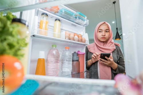 asian muslim young woman using her smartphone to buy groceries while open her fridge at home