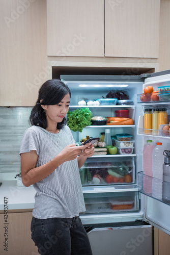 asian young woman using her smartphone to buy groceries while open her fridge at home