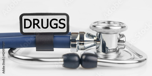 On the white surface lies a stethoscope with a plate with the inscription - DRUGS