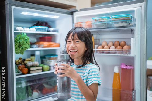 thirsty happy young asian girl open fridge door drinking a bottle of water