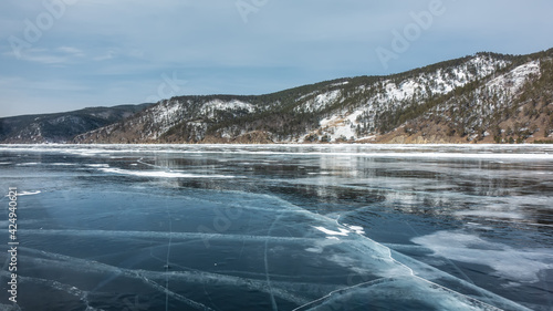 The smooth shiny ice of the lake is streaked with deep cracks. On the shore there are snow-capped wooded mountains. Reflection on the surface. Baikal in winter