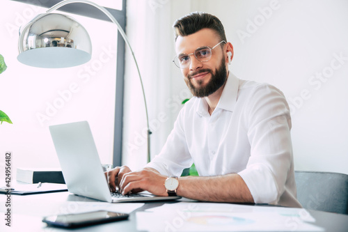 Young modern handsome stylish bearded businessman in glasses and a white shirt is working on the laptop with documents, papers on the desk and looking on the camera.