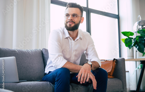 Confident stylish handsome bearded business man in glasses and formal smart wear is sitting on the couch in the modern office or room