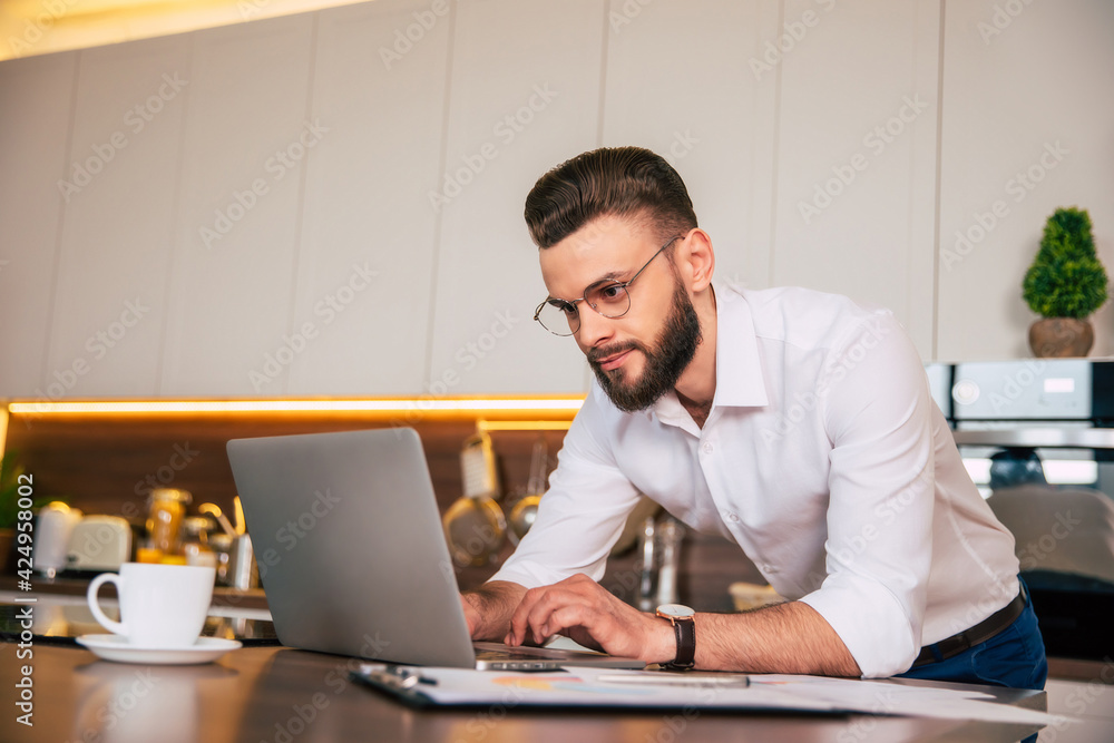 Handsome confident businessman in smart casual wear is working with laptop at domestic kitchen Handsome confident businessman in smart casual wear is working with laptop at domestic kitchen