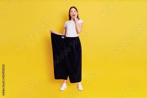 Caucasian shocked female with dark hair looking at camera with big eyes and open mouth, covering cheek with palm, wearing too big pants, woman lost weight, isolated over yellow background. photo