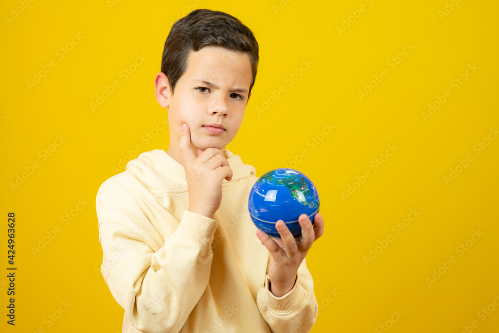 Pensive young caucasian boy holding a world globe over yellow background Pensive young caucasian boy holding a world globe over yellow background