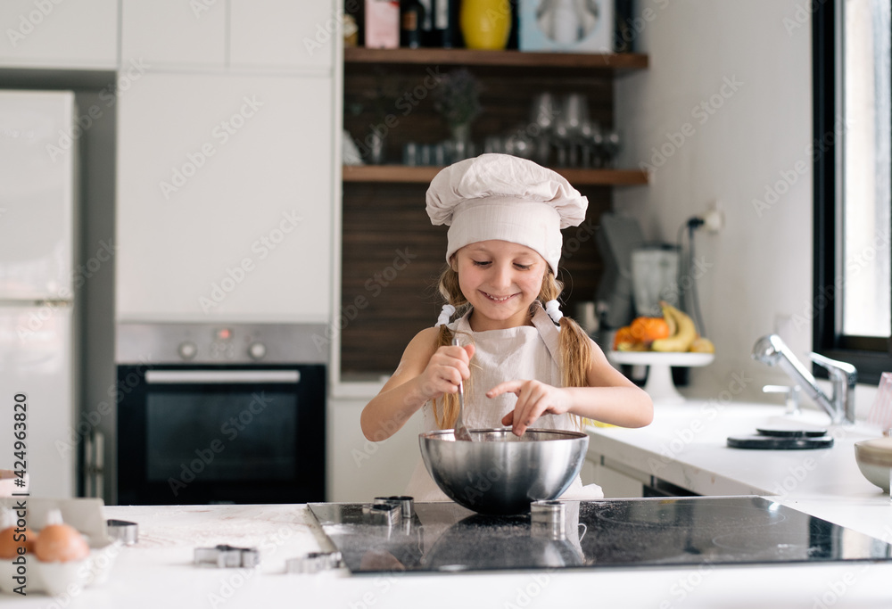 Small cute girl in special clothes and funny chef cap stay on kitchen and make dough for cake. Making cookie at home on kitchen, kid get fun enjoy cook. Child make dessert Small cute girl in special clothes and funny chef cap stay on kitchen and make dough for cake. Making cookie at home on kitchen, kid get fun enjoy cook. Child make dessert