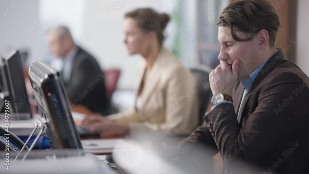 Tired handsome overworked man yawning typing on computer keyboard with busy blurred coworkers working online at background. Exhausted Caucasian employee at workplace in office Tired handsome overworked man yawning typing on computer keyboard with busy blurred coworkers working online at background. Exhausted Caucasian employee at workplace in office