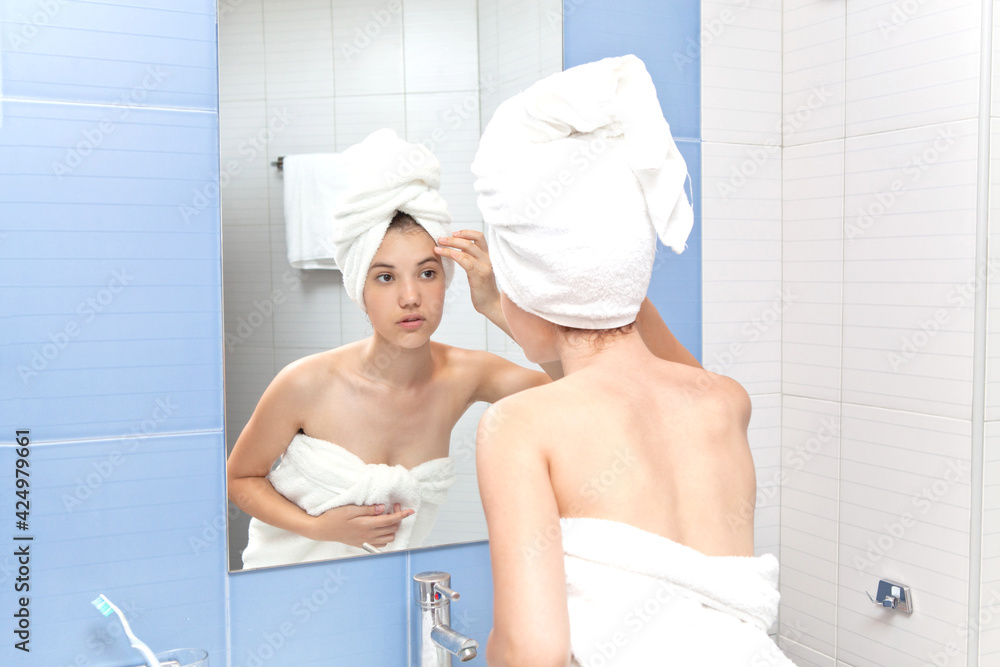A young woman looks at her face while standing in front of a mirror in a home bathroom. Beautiful girl in a bath towel in the head portrait. Skin care after cleansing A young woman looks at her face while standing in front of a mirror in a home bathroom. Beautiful girl in a bath towel in the head portrait. Skin care after cleansing