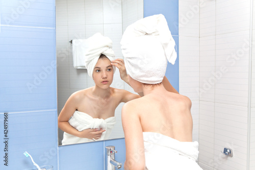 A young woman looks at her face while standing in front of a mirror in a home bathroom. Beautiful girl in a bath towel in the head portrait. Skin care after cleansing