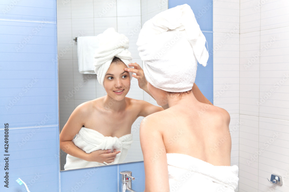 A beautiful young girl in a white bath towel on her head and body in the bathroom looks at her face. Hygiene A beautiful young girl in a white bath towel on her head and body in the bathroom looks at her face. Hygiene