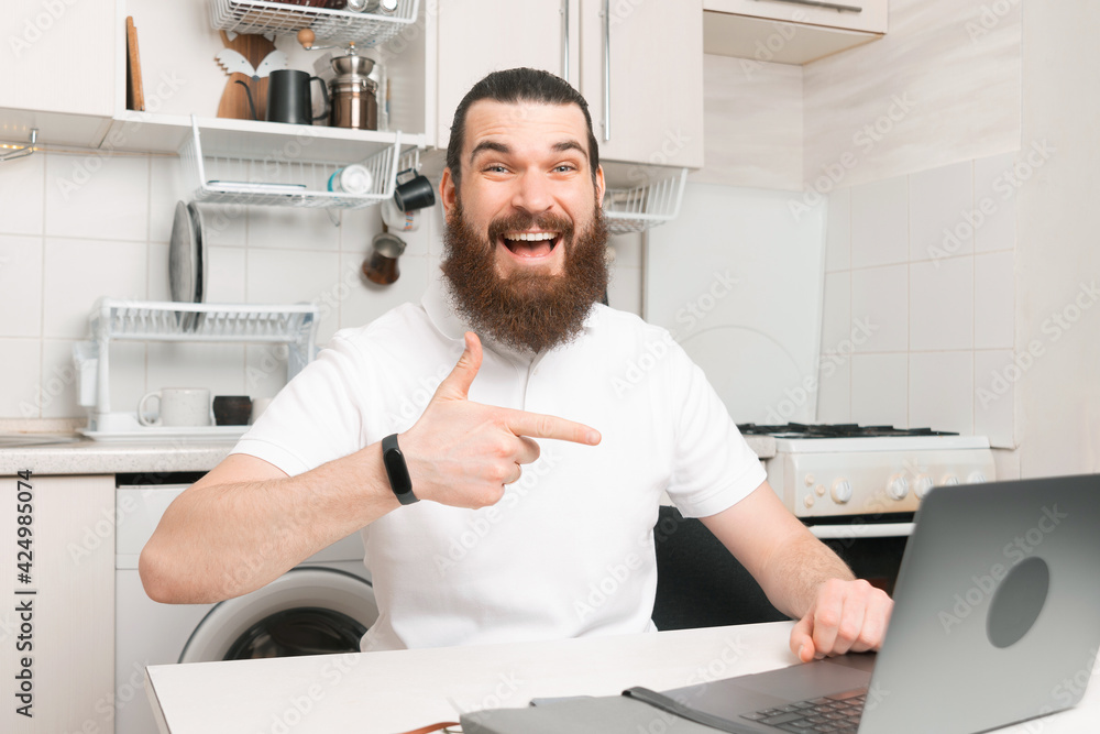 Happy amazed young bearded hipster man sitting in kitchen and pointing at laptop, work from home Happy amazed young bearded hipster man sitting in kitchen and pointing at laptop, work from home