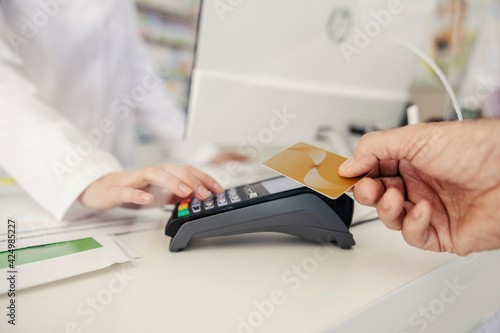 Payment by card at the pharmacy. Male hands provide a payment credit or debit card to the terminal while female hands of the pharmacist enter the pin code. Purchase of drugs for therapy