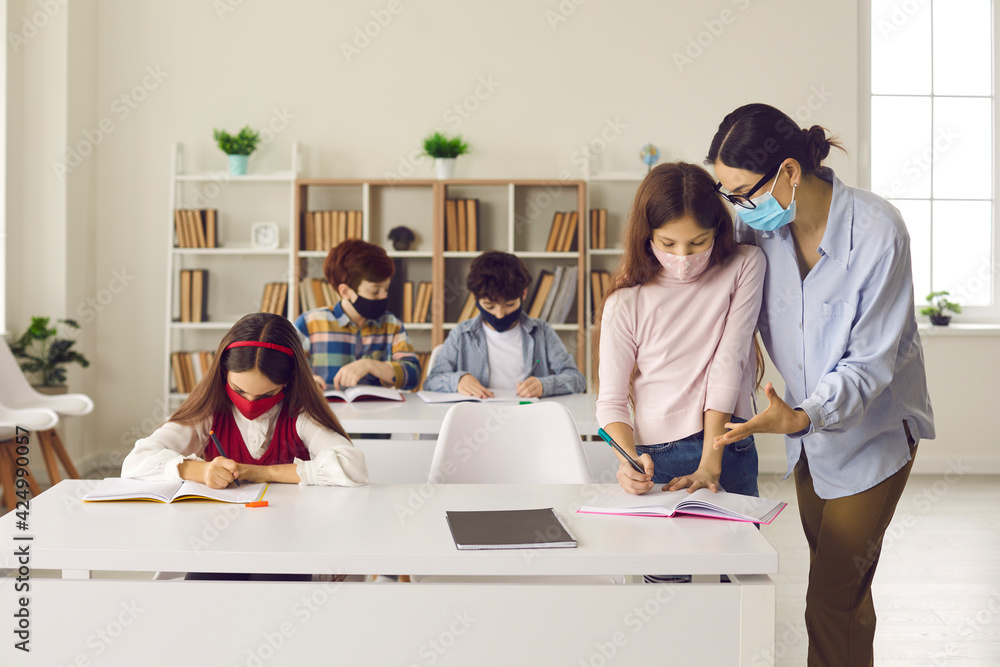 Teaching and pandemic. Teacher and children with face mask back at school after covid-19 quarantine and lockdown. Female teacher helps a little girl student in a class with a small number of students. Teaching and pandemic. Teacher and children with face mask back at school after covid-19 quarantine and lockdown. Female teacher helps a little girl student in a class with a small number of students.