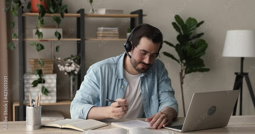 Man sit at desk wear headset studying with online tutor, talk to teacher e learning foreign language using laptop and video call app, gain knowledge, makes notes. Distance education, e-learn concept Man sit at desk wear headset studying with online tutor, talk to teacher e learning foreign language using laptop and video call app, gain knowledge, makes notes. Distance education, e-learn concept